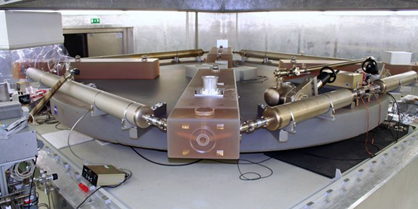 In a laboratory, metal tubes are arranged in a square pattern on a circular disc. There are also cables, control units and measuring instruments.