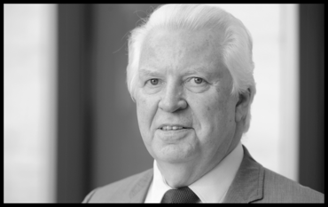 A black-and-white portrait photograph. An elderly man with white hair, wearing a suit and a dark tie, looks friendly and directly into the camera.