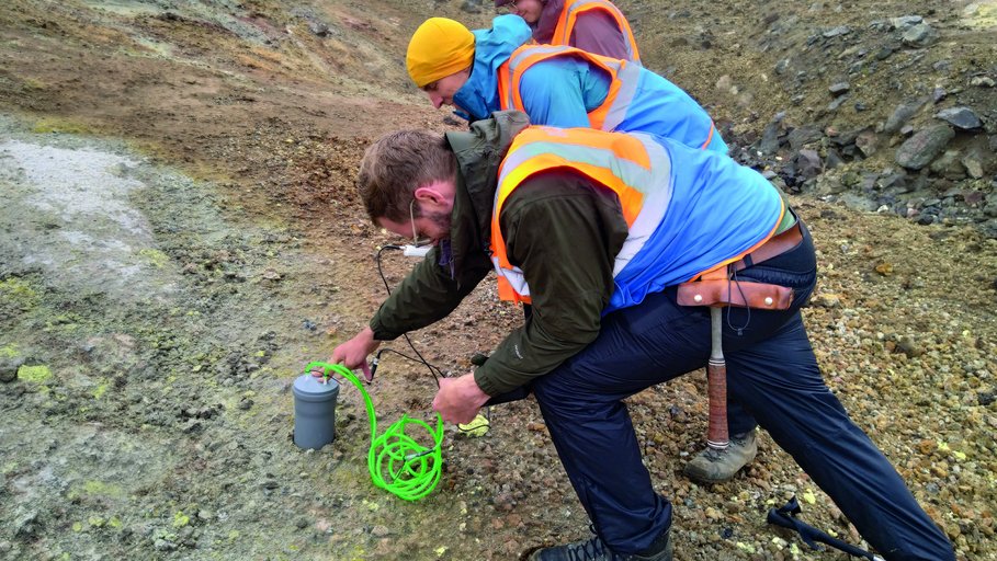 Temperature and multi-gas flow measurements on a caldera. Three people in blue and orange vests are working along the caldera, bent over to take measurements.