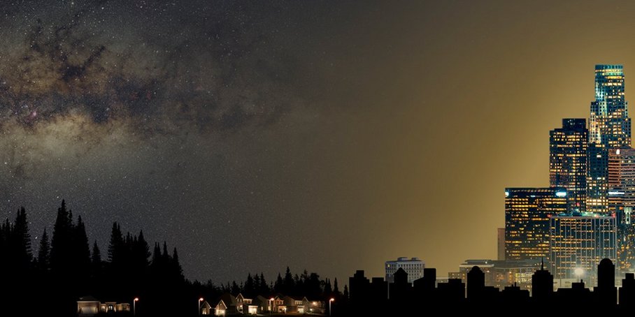 A full starry sky with Milky Way can be seen above sparsely built-up terrain. Towards the left, the development - and thus the artificial lighting - increases more and more, and at the same time fewer and fewer stars become visible in the night sky.