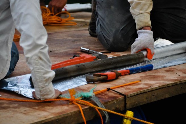 Auf einem Brett wird ein Sedimentbohrkern aus einer Metallhülle geschoben. Daneben Werkzeug.