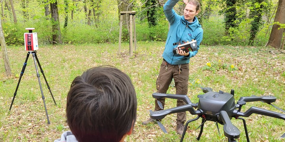 The image shows a student in a red jacket from behind in a meadow in the forest, watching a man in a blue jacket and field attire explain the spider-like drone standing next to him.