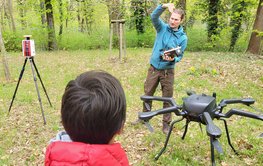 The image shows a student in a red jacket from behind in a meadow in the forest, watching a man in a blue jacket and field attire explain the spider-like drone standing next to him.