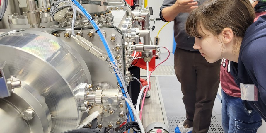 Eine Schülerin, rechts im Bild, schaut gebeugt auf eine silberne Maschine, aus der blaue Kabel führen. Hinter ihr steht ein Wissenschaftler, dessen Kopf vom Bild abgeschnitten ist und etwas erklärt.