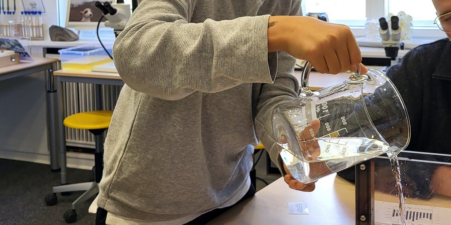 Drei Schülerinnen stehen von hinten fotografiert vor einem Setzkasten mit Sand gefüllt, welcher Flusssysteme nachstellt.