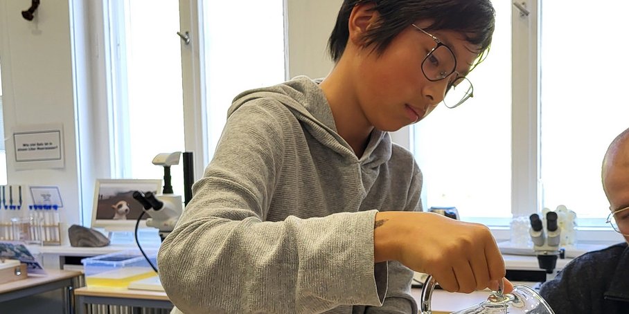 A student is holding a glass jug and, leaning over, is pouring a clear liquid into a glass trough that is cut off from the photo. In front of him on the table is a tray with measuring tools and small glasses.