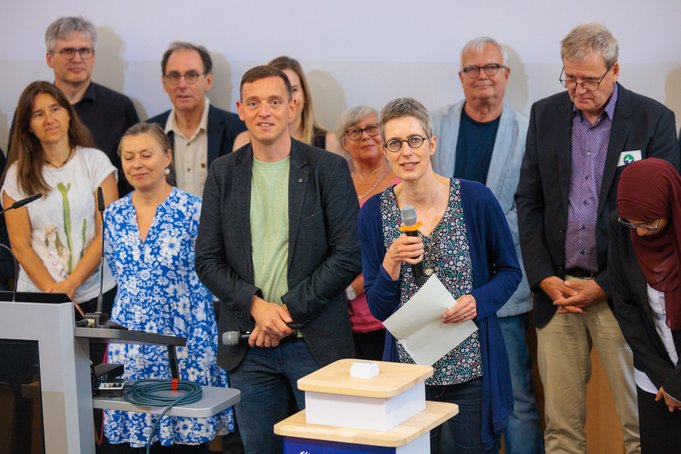 Close-up of a group of 10 people. Two people are standing at the front with microphones at a small lectern; a woman with glasses is speaking.