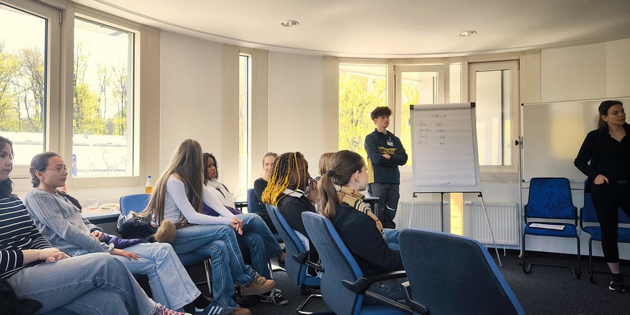 Schülerinnen und Schüler sitzen in einem runden Sitzungsraum links und schauen auf einen Vortrag rechts. Die Wand ist mit Fenstern übersehen und weiß.