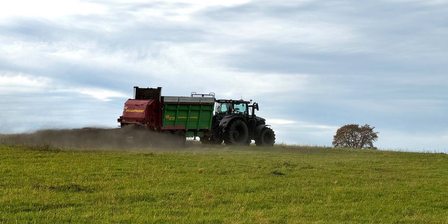 A tractor with a trailer spraying finely ground basalt drives across a field of grass.