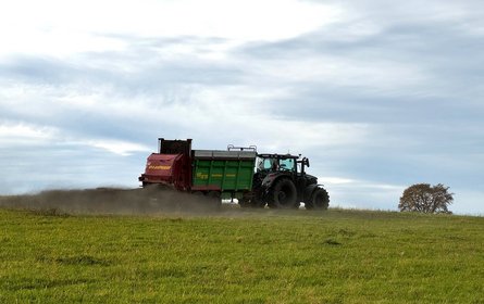 A tractor with a trailer spraying finely ground basalt drives across a field of grass.