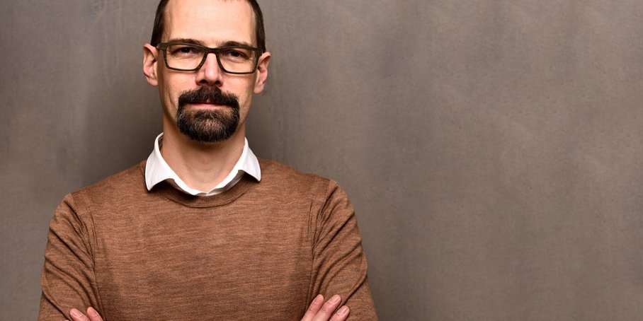 Sascha Brune, man with brown hair, glasses and beard in brown jumper with white collar, brown background
