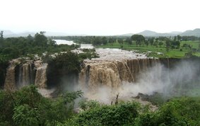 Wide waterfalls in a green landscape with grass and trees. Far: middle high mountains.
