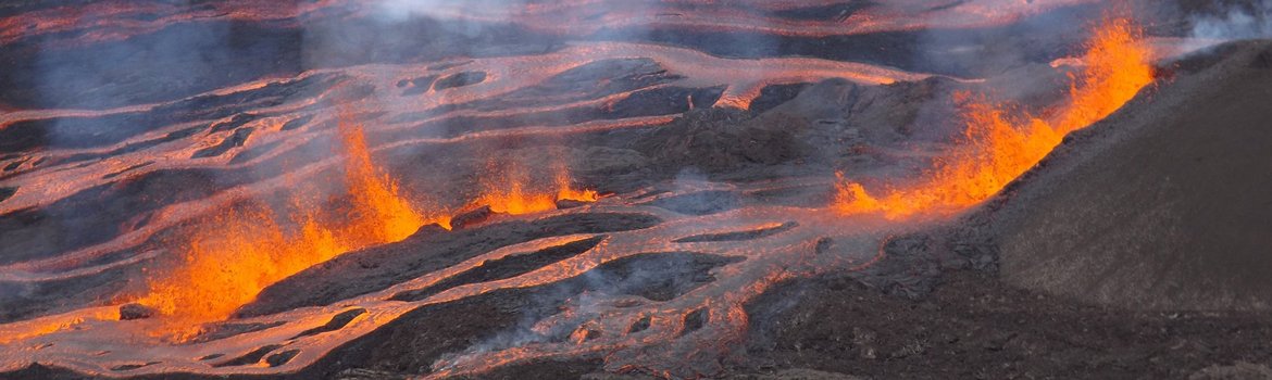 Verschlungene rote Lavaströme auf schwarzem Untergrund. Im Vordergrund spritzende rote Lava, in der Luft Rauchschwaden.