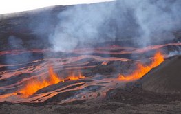 Verschlungene rote Lavaströme auf schwarzem Untergrund. Im Vordergrund spritzende rote Lava, in der Luft Rauchschwaden.