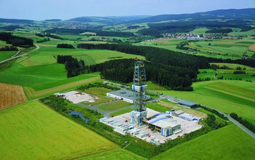 A square drilling tower made of metal rods, 83 metres high, surrounded by flat buildings in a hilly green landscape.