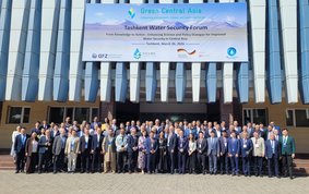 Group photo of around 100 people in front of a building displaying a large poster announcing the event.