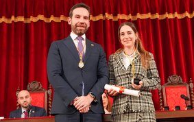 Eine junge Frau mit dunklen langen Haaren und ein Mann stehen vor einem samtroten Vorhang. Sie trägt eine goldene Medaille um den Hals und eine Urkunde in der Hand.