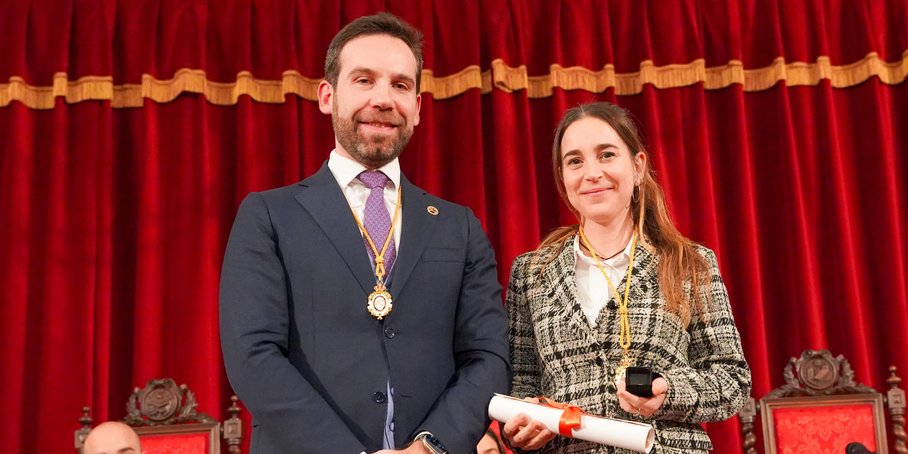 A young woman with long dark hair and a man are standing in front of a velvet red curtain. She is wearing a gold medal around her neck and holding a certificate in her hand.