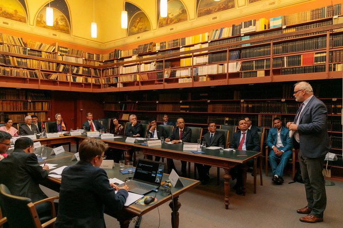 A white-haired man stands in the library and speaks. The audience is seated around tables in front of him.