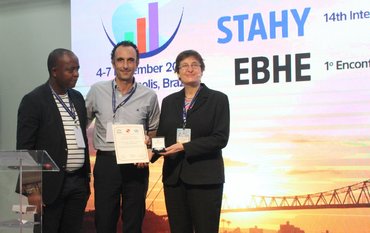 The awardee stands to the right of two men and holds a certificate and a medal. In the background is a screen with a sunset and the conference logos.