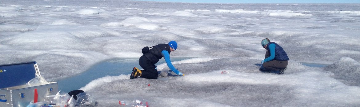 On a vast expanse of white-gray ice and snow dotted with light blue pools of meltwater, two people kneel and collect snow samples in small plastic tubes. In front of them is an open silver suitcase containing equipment, some of which is lying on the ice.