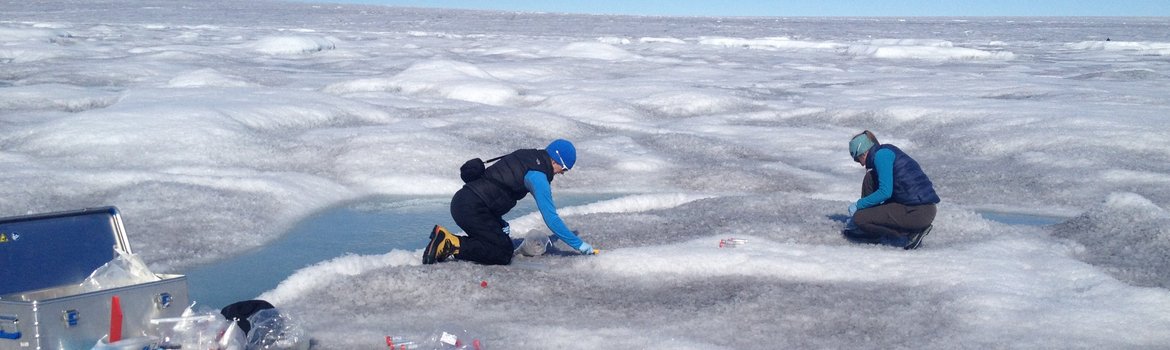 Auf einer riesigen weiß-grauen Eis- und Schneefläche mit hellblauen Schmelzwasserpfützen knien zwei Personen und sammeln Proben. Vorne ein silberner Koffer mit Equipment.