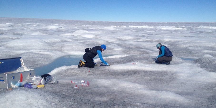 On a vast expanse of white-gray ice and snow dotted with light blue pools of meltwater, two people kneel and collect snow samples in small plastic tubes. In front of them is an open silver suitcase containing equipment, some of which is lying on the ice.