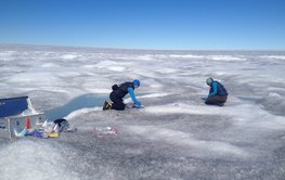 Auf einer riesigen weiß-grauen Eis- und Schneefläche mit hellblauen Schmelzwasserpfützen knien zwei Personen und sammeln Proben. Vorne ein silberner Koffer mit Equipment.