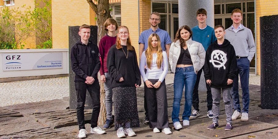 Nine of the new trainees, dual students and FJN students in front of the pillar forum with the GFZ logo in the background.