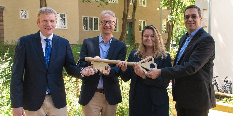 Vier Personen halten einen großen goldenen Schlüssel vor einem Wohnhaus