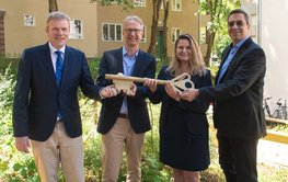 Vier Personen halten einen großen goldenen Schlüssel vor einem Wohnhaus