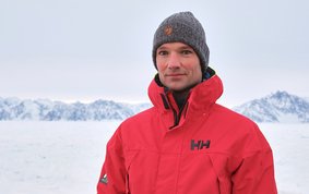 Tim Kalvelage, wearing a red functional jacket, sits in front of a snow-covered white background, with snow-capped mountains on the horizon and just a few rocks jutting out into the sky.
