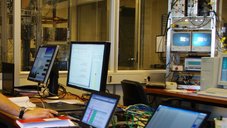 The photo shows the control room of a Cyclotron radiation source during a GNSS receiver test. On a table there are 3 computer displays which monitor the function of the GNSS-receiver under influence of strong radiation is monitored, using a specially adapted graphical user interface.
