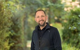 Porträt eines Mannes vor unscharfen grünen Büschen: Der Mann trägt dunkle Kleidung, einen Bart und lächelt freundlich.