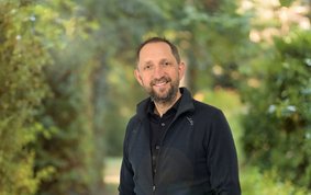 Porträt eines Mannes vor unscharfen grünen Büschen: Der Mann trägt dunkle Kleidung, einen Bart und lächelt freundlich.