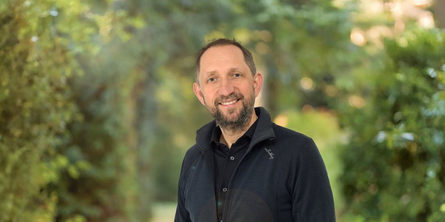 Portrait of a man in front of blurred green bushes: The man is wearing dark clothes, has a beard and is smiling warmly.