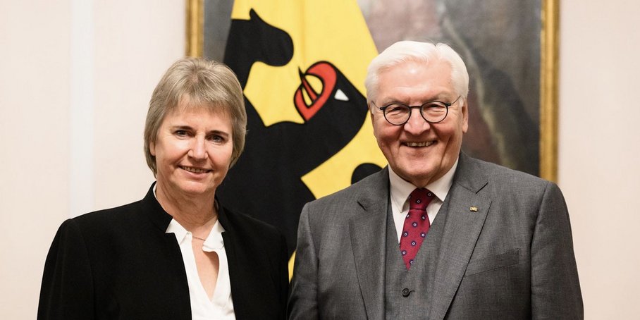 Portrait of two friendly smiling persons in front of a German Flag and a historic painting: A mid-aged woman with blond short hair and a black suit on the left, a white-haired man wearing a grey suit and glasses on the right.