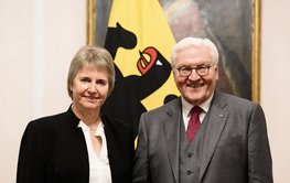 Porträt zweier freundlich lächelnder Personen vor einer deutschen Flagge und einem historischen Gemälde: Links eine Frau mittleren Alters mit blondem Kurzhaarschnitt und schwarzem Anzug, rechts ein weißhaariger Mann mit grauem Anzug und Brille.