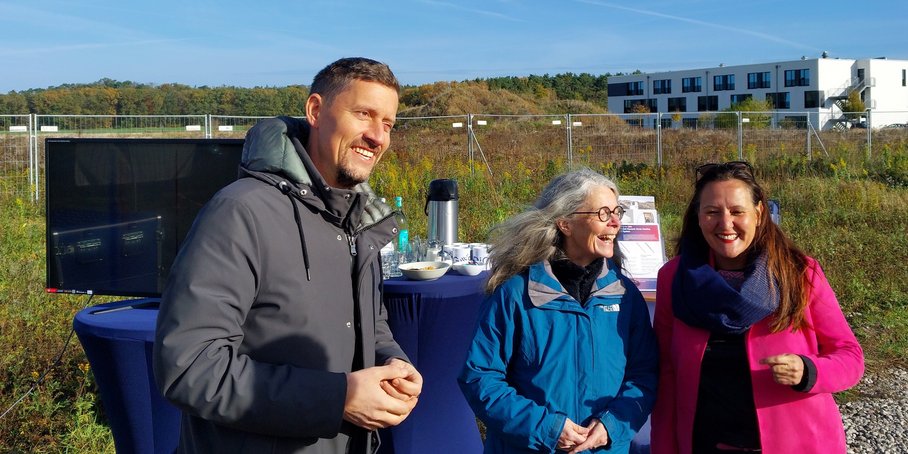 Drei Personen stehen in freiem Gelände. Im Hintergrund ein Messgerät, Stehtische, ein Bauzaun und Baucontainer.