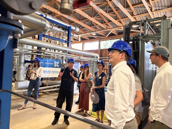 Eine Gruppe von Menschen mit Schutzhelm steht in einer Halle mit technischen Rohren und Anlagen.