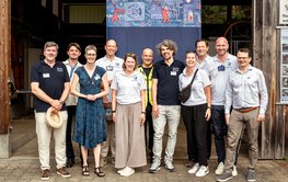 Group photo of eleven people in front of the entrance gate to a hall. Behind them hangs a large poster with a colourful image.