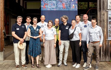 Group photo of eleven people in front of the entrance gate to a hall. Behind them hangs a large poster with a colourful image.