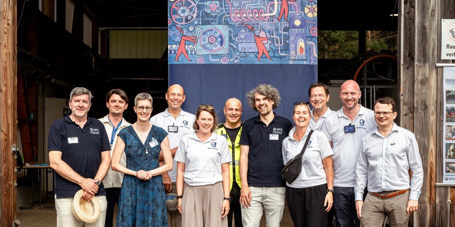 Gruppenfoto mit elf Personen vor dem Eingangstor zu einer Halle. Dahinter hängt ein großes Plakat mit buntem Bild.