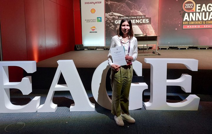 Junge Frau steht mit Urkunde in der Hand in einer Halle vor dem Schriftzug EAGE
