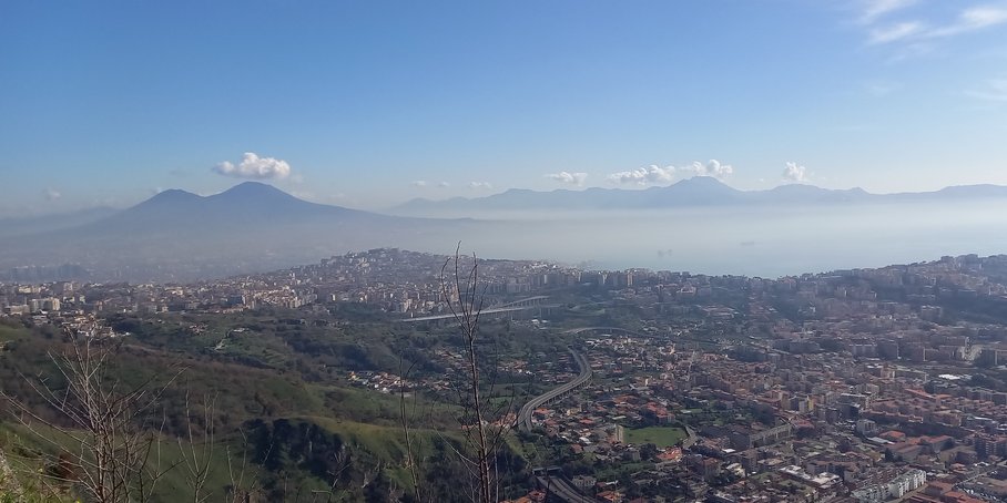 Urban area at the foot of a mountain, with further mountains and the sea in the background.