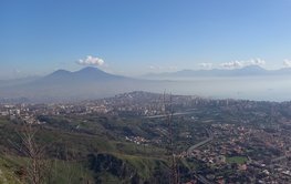 Urban area at the foot of a mountain, with further mountains and the sea in the background.