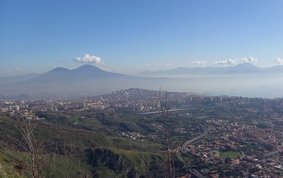 Urban area at the foot of a mountain, with further mountains and the sea in the background.
