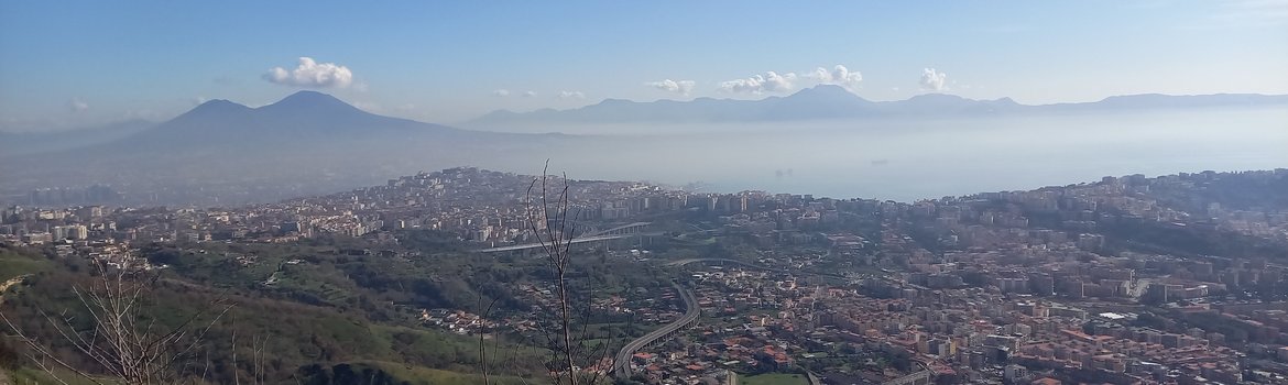Urban area at the foot of a mountain, with further mountains and the sea in the background.