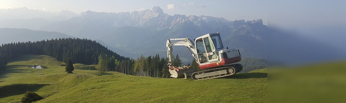 Auf dem Bild ist eine grüne Wiese im Vordergrund und eine blaue Alpenlandschaft im Hintergrund zu sehen, rechts mittig steht ein weißer Bagger.