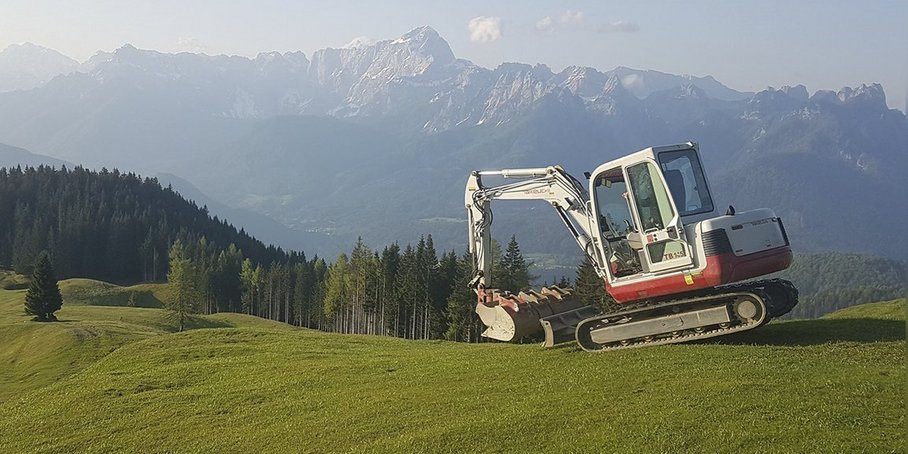 Auf dem Bild ist eine grüne Wiese im Vordergrund und eine blaue Alpenlandschaft im Hintergrund zu sehen, rechts mittig steht ein weißer Bagger.